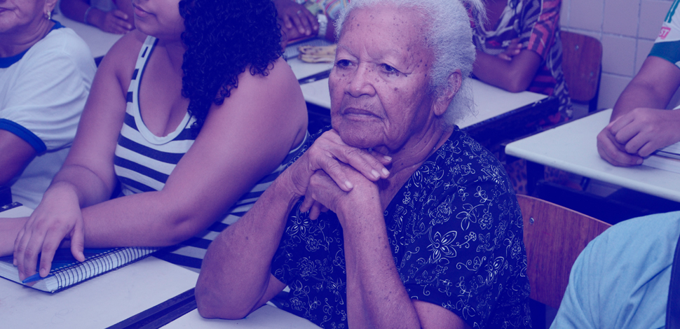 Idosa sentada em sala de aula, na companhia de outros adultos.
